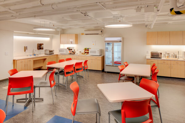 Tables and bold red chairs in a break room with a kitchen