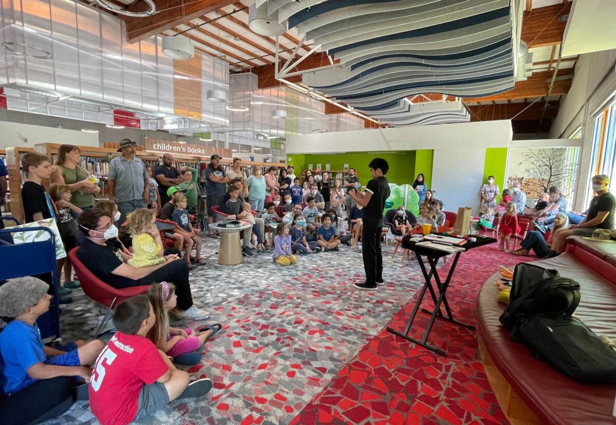 A young performer in front of a rapt audience of all ages gathered in a semicircle in the renovated children's area with tall ceilings and striking wavy acoustic panels